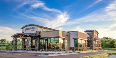 A modern bank with large windows and stone accents under a blue sky with wispy clouds. The foreground features a landscaped area with grass and a paved parking lot.