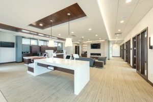 A modern design-build office space with a long white desk featuring built-in outlets and two white chairs. The room includes dark wood accents, seating areas with couches, multiple doorways lined along one wall, and a wall-mounted TV. Large windows bring in natural light.