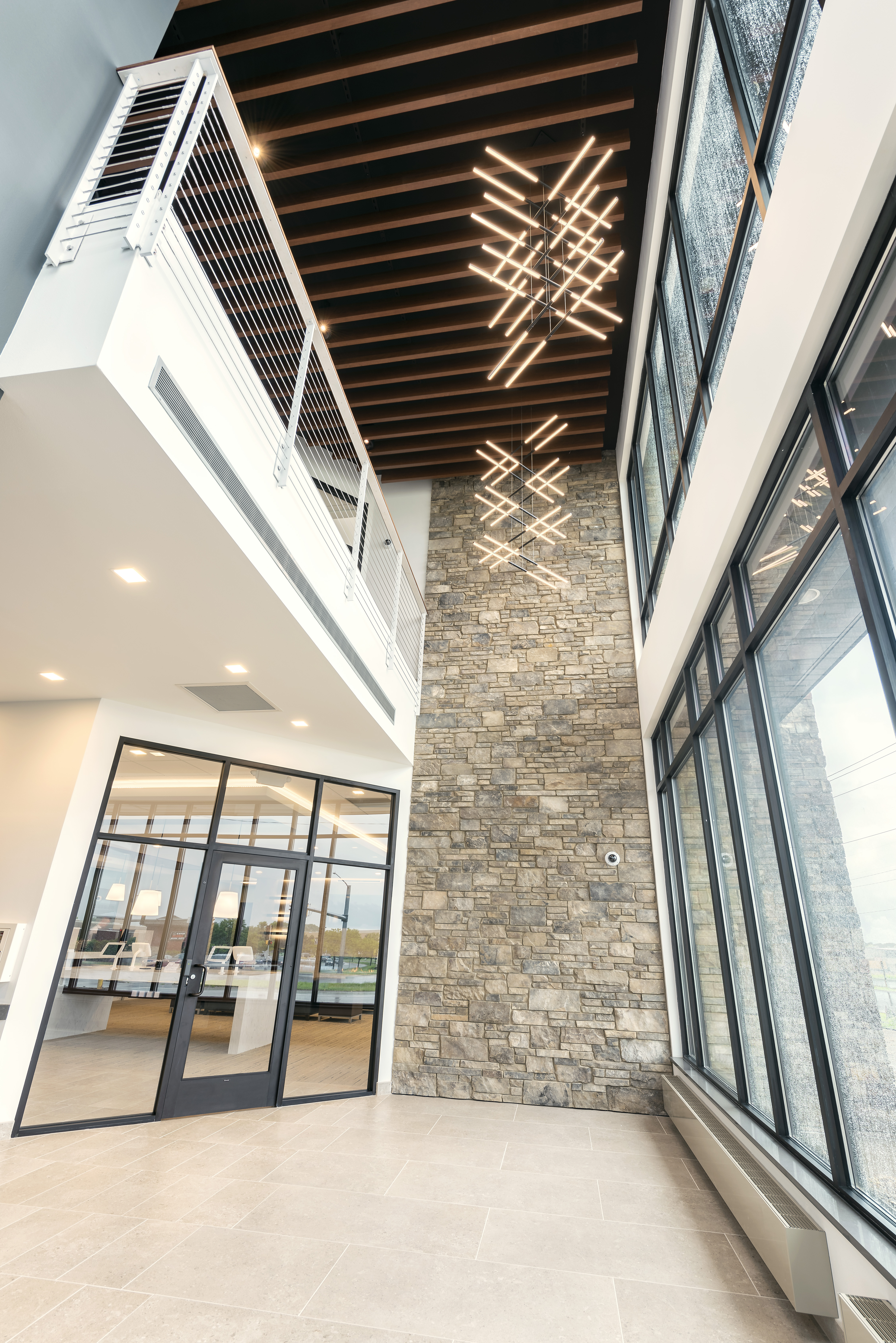 A modern, high-ceilinged lobby with a stone accent wall and large windows, allowing natural light to illuminate the space. Contemporary, geometric light fixtures hang from the wood-beam ceiling above a glass door entrance.