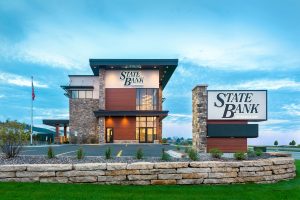 A modern, two-story building with "State Bank" signs, featuring stone accents and large windows. It stands in a landscaped area with a stone retaining wall and a grassy lawn.