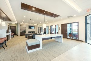 A modern design-build office space with a long white desk featuring built-in outlets and two white chairs. The room includes dark wood accents, seating areas with couches, multiple doorways lined along one wall, and a wall-mounted TV