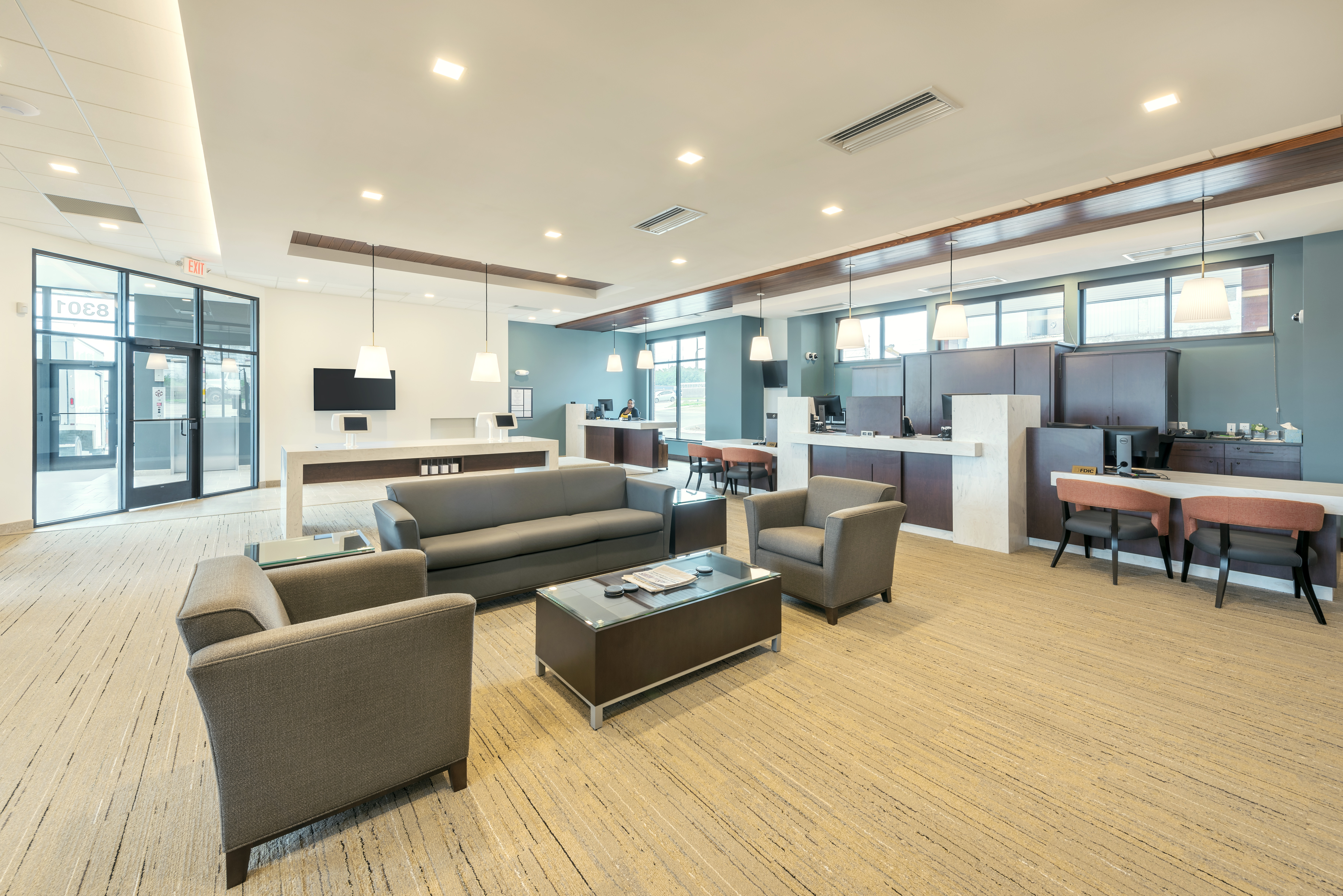 A modern office reception area features a seating arrangement with gray armchairs and a sofa around a coffee table. The space includes workstations with computers