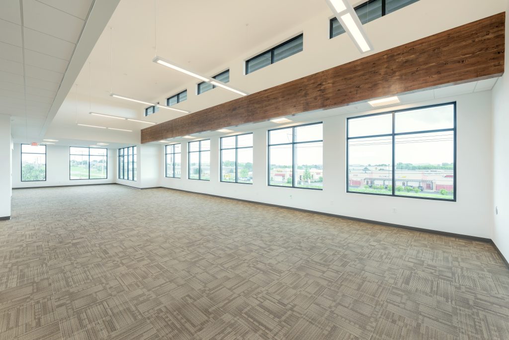 A spacious, empty office with large windows providing ample natural light. The room features a mix of carpeted flooring and exposed wooden beams on the ceiling.