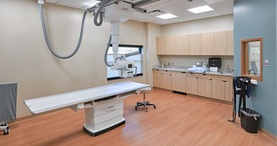 A sterile, well-lit medical examination room with a large imaging machine and table in the center. Cabinets, a sink, and countertop fill the back wall. A window lets in natural light, and a small stool is beside the table. Floor is wooden, and walls are painted light.