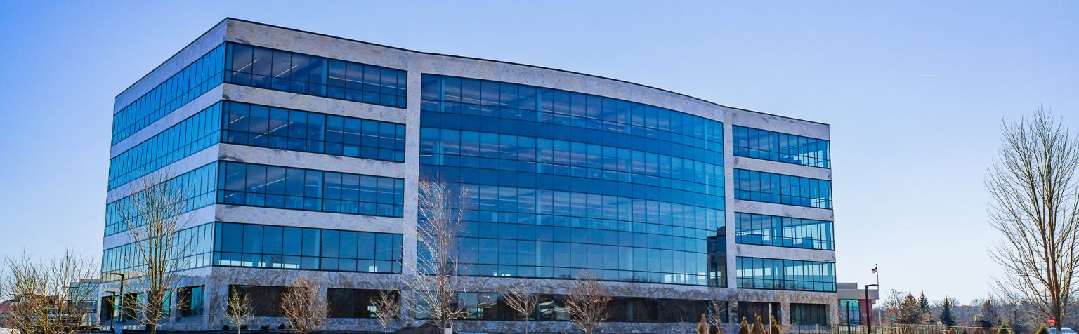 glass and stone office building on a clear day. The building has large, reflective glass windows and a sleek facade. The surrounding area includes a construction site and small landscaped trees.