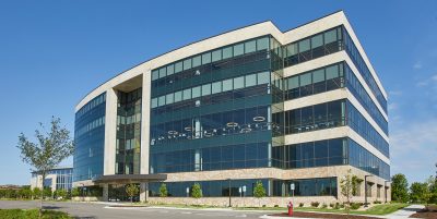 glass and stone office building on a clear day. The building has large, reflective glass windows and a sleek facade. The surrounding area includes a construction site and small landscaped trees.