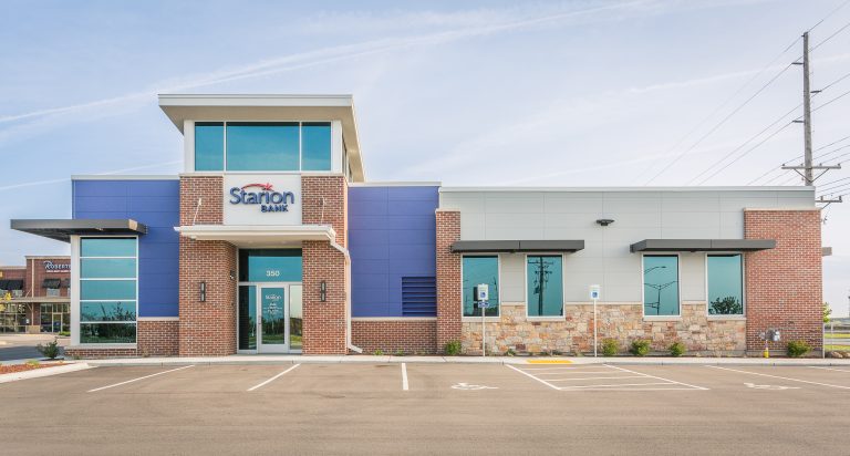 A modern bank building with a mix of brick, glass, and stone exterior. The entrance features large glass doors, and an accessible parking spot is in the foreground.