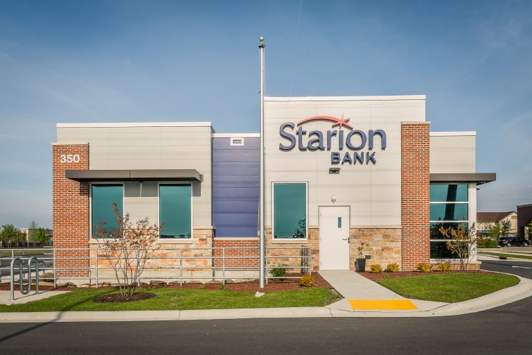 Exterior bank building with a brick and metal facade. The building is surrounded by a clean sidewalk, newly planted trees, and a small parking lot.