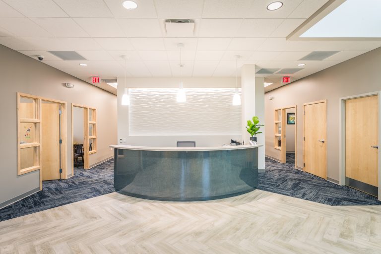 A modern reception area with a semi-circular desk and a textured white wall behind it. The space is well-lit with hanging pendant lights. There are wooden doors on either side and blue patterned carpet with light-colored flooring.