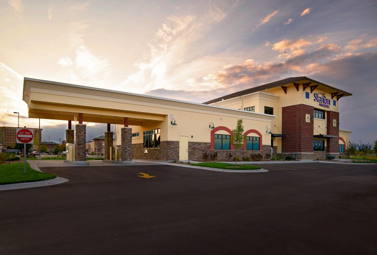 A modern bank at sunset features tan and brick exteriors, large windows, and a covered drive-thru area. The sky is partly cloudy with warm light.