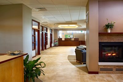 A clean, modern reception area features a wooden desk, potted plants on either side, and neutral-colored walls. Overhead, there is a circular light fixture set in a recessed ceiling. Glass-paneled doors are visible in the background.