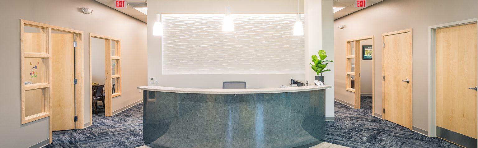 A modern reception area with a semi-circular desk and a textured white wall behind it. The space is well-lit with hanging pendant lights.