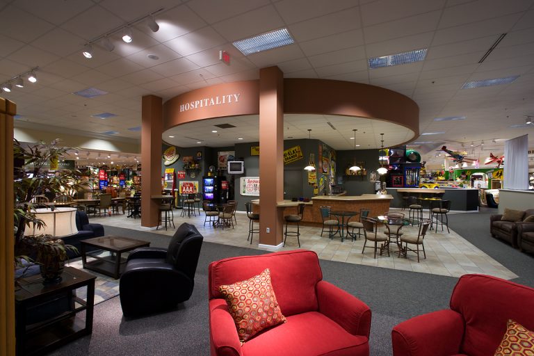 A spacious, well-lit hospitality area with red armchairs in the foreground. There are several tables and chairs arranged in the middle. The room extends into a larger area filled with arcade games and decorative elements.