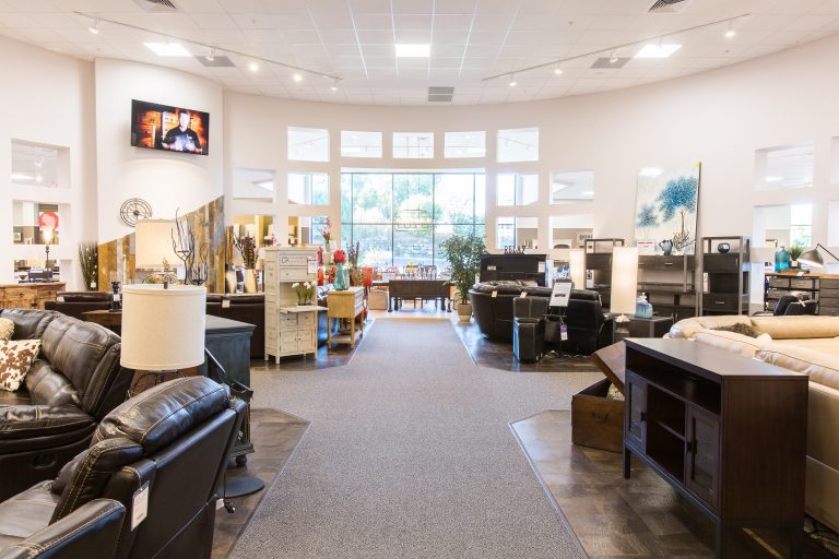 A furniture store showroom featuring various items like sofas, tables, and cabinets. The space is well-lit with large windows at the back. A TV is mounted on the wall, and decor pieces are arranged throughout the store.