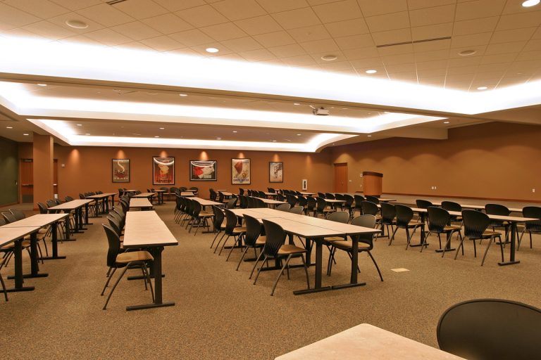 A spacious conference room with rows of long tables and black chairs arranged classroom-style. The ceiling has recessed lighting, and several artworks hang on a brown wall. A podium stands in the front right corner.