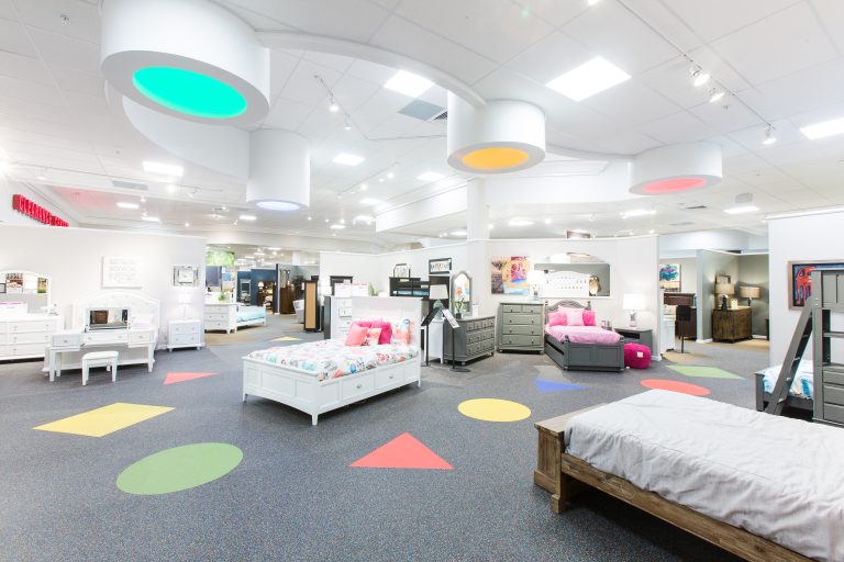 A spacious furniture showroom. The room features colorful, circular lights on the ceiling and geometric shapes on the carpet. Beds, dressers, and decor items are arranged throughout the space.