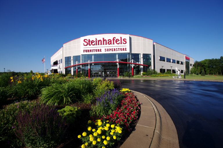 A large corporate, retail building with a sign that reads "Steinhafels Furniture Superstore" is surrounded by colorful flowers and manicured gardens. The sky is clear and blue, and there is a curved driveway leading to the entrance.