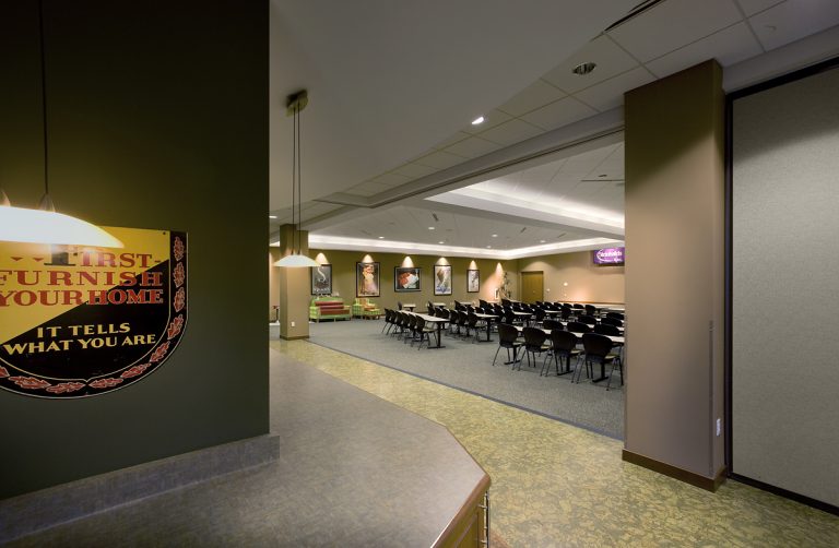 A spacious conference room with rows of black chairs facing a stage. The room has green and beige walls with posters. Ceiling lights are evenly spaced, and there's a signage board on the left wall.