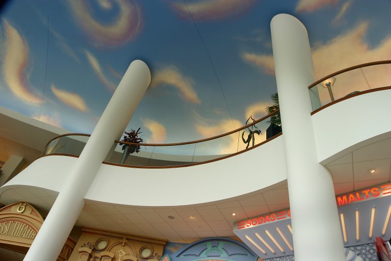 Interior of a lobby with a painted sky ceiling featuring swirling cloud patterns. White columns and a curved railing surround the upper level.