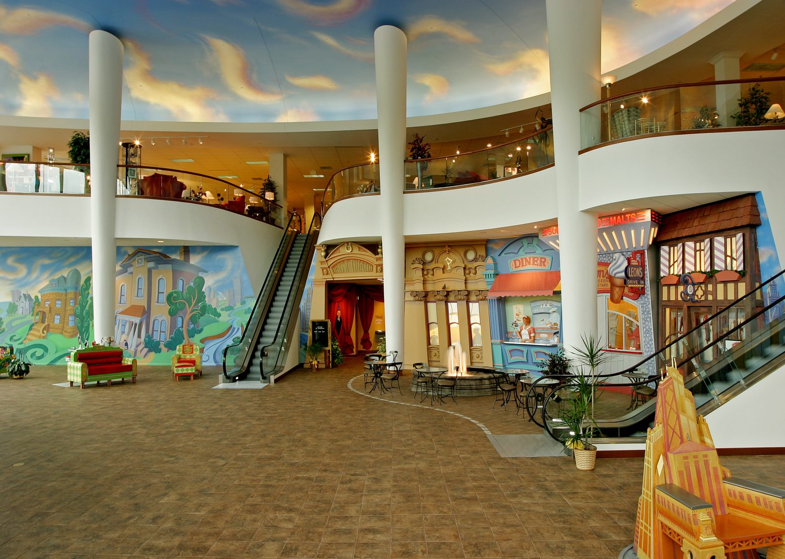 A colorful indoor shopping center with a large open space features a decorative mural depicting a diner and buildings. An escalator leads to an upper level.