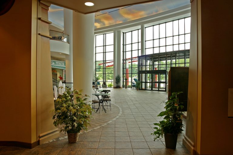 Bright lobby with large windows and a high ceiling. The floor is tiled, and there are potted plants along the walls. A few tables and chairs are arranged near a small café in the background.