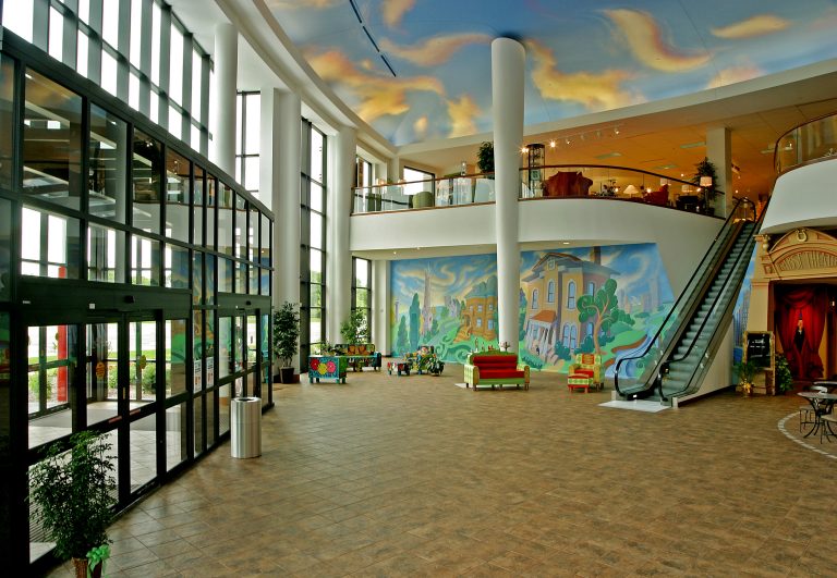 Spacious, modern lobby with large windows, a colorful mural on the wall, and an escalator leading to the second floor. The ceiling features a cloud design, and furniture includes couches and a small coffee table.