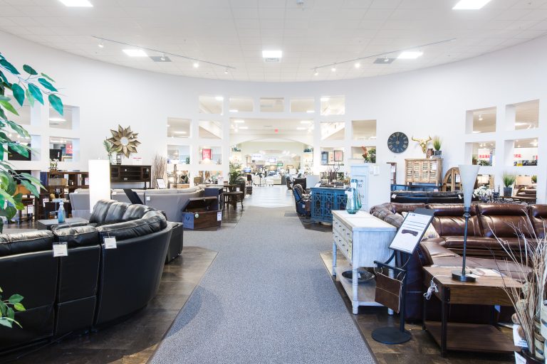 A spacious furniture store with various sofas and tables on display. The interior features a high ceiling, neutral walls, and a carpeted walkway. Decorative items, plants, and wall clocks are visible throughout the showroom.