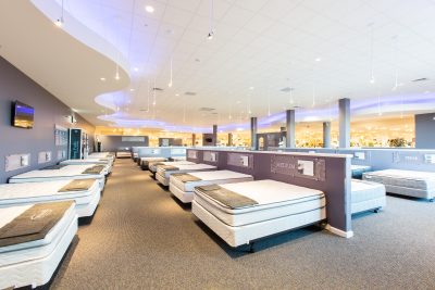 A spacious showroom featuring neatly arranged mattresses on display. The room has modern lighting with a wavy ceiling design. Various mattress models are lined up in rows, each with pillows and decorative elements atop them.