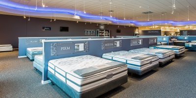 showroom featuring neatly arranged mattresses on display. The room has modern lighting with a wavy ceiling design.