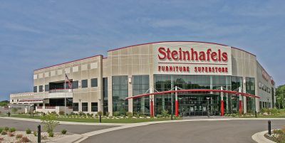 A large retail building with a sign that reads "Steinhafels Furniture Superstore" is surrounded by colorful flowers and manicured gardens. The sky is clear and blue, and there is a curved driveway leading to the entrance.