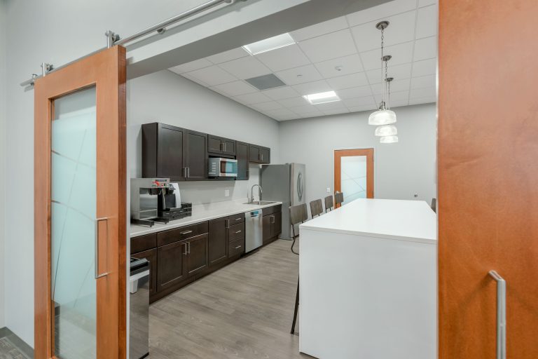 Office kitchen with dark wood cabinets, stainless steel appliances, and a light wood floor. There is a white island in the center with bar stools. Pendant lights hang from the ceiling. The room is accessed through sliding glass doors with wood frames.