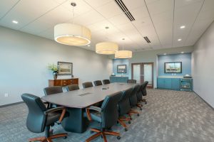 A spacious conference room with a long table surrounded by ten black leather office chairs. The room is well-lit by three large pendant lights hanging from the ceiling. Artworks and cabinets are positioned against the back wall. The floor is carpeted in a gray pattern.