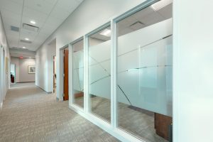 A modern office corridor with light beige carpeting and a white ceiling featuring recessed lighting. The right side of the hallway has large windows with frosted glass panels and an abstract pattern. At the end of the corridor, there is a wooden door and wall art.