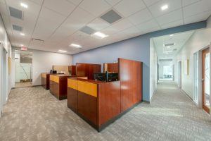 A modern, brightly-lit office interior with a hallway leading to an open area. The office has wooden cubicles with computers on desks. The floor is carpeted, and the walls are painted in light, neutral colors. Recessed lighting and ceiling tiles are present.