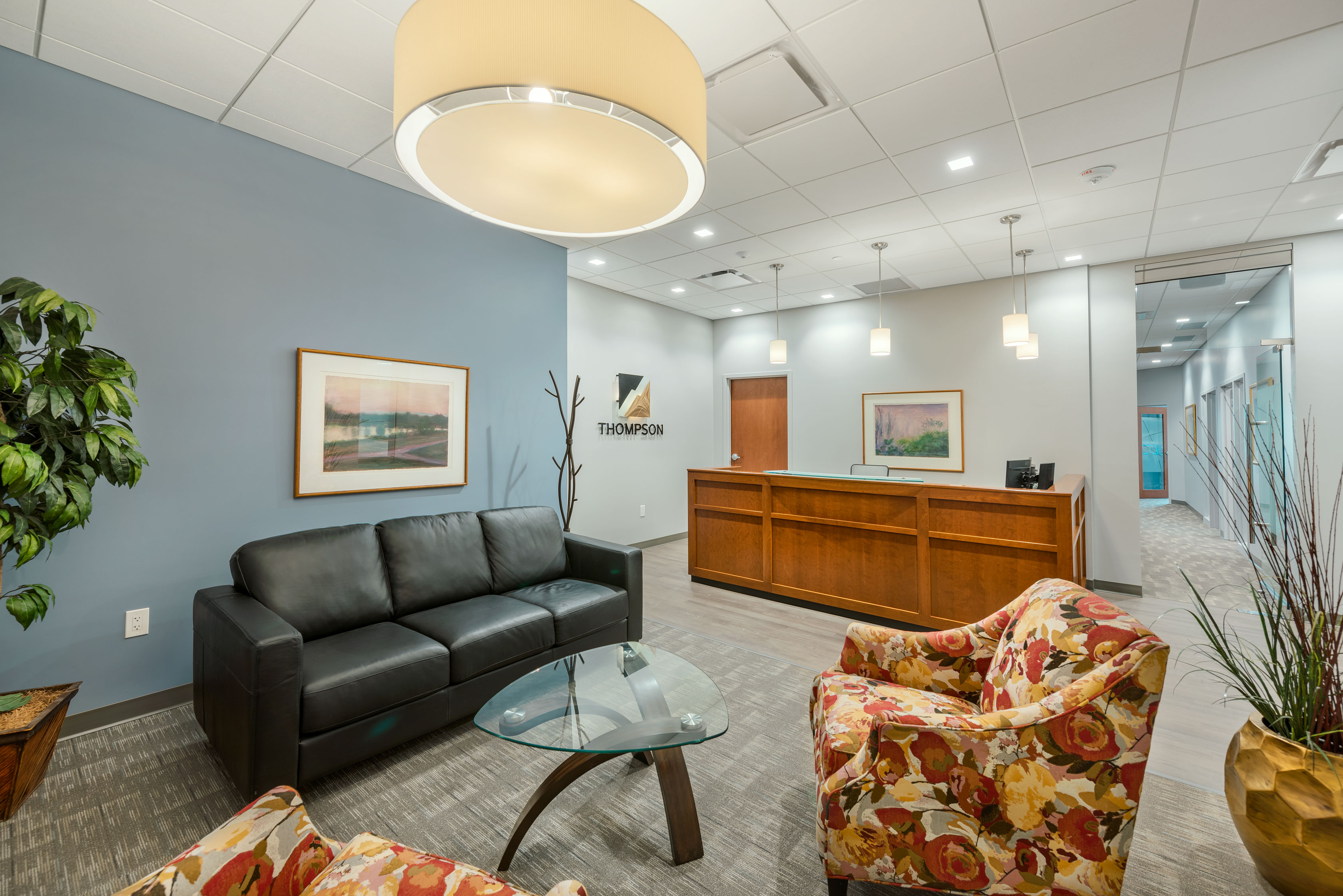 office reception area with a black leather couch, two floral armchairs, and a glass coffee table. A wooden reception desk is in the background beneath a sign with the word "THOMPSON." The ambiance is enhanced by various artworks and plants.