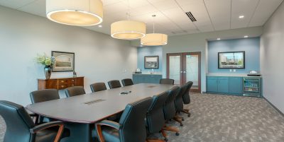 A spacious conference room with a long table surrounded by ten black leather office chairs. The room is well-lit by three large pendant lights hanging from the ceiling. Artworks and cabinets are positioned against the back wall. The floor is carpeted in a gray pattern.