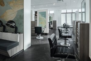 A modern and stylish hair salon interior featuring sleek black leather styling chairs, hairdryers, wood-textured counters, and large mirrors. Soft natural light filters in through large windows. Decor includes a subtle leafy mural on one wall.