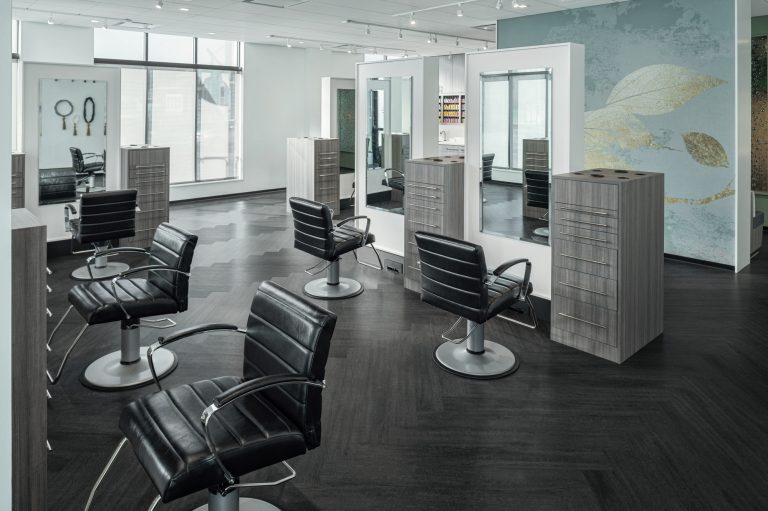 hair salon with multiple black leather chairs and large mirrors along a wall. The room is well-lit, featuring a wooden floor with a dark finish and decorative abstract artwork on the walls. Tall windows allow natural light to fill the space.