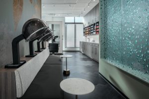 A modern hair salon with drying stations on the left, featuring dome-shaped dryers above a padded bench. To the right, there's a wall of nail polish shelves and washing stations. The room has sleek, contemporary decor with a textured glass partition.