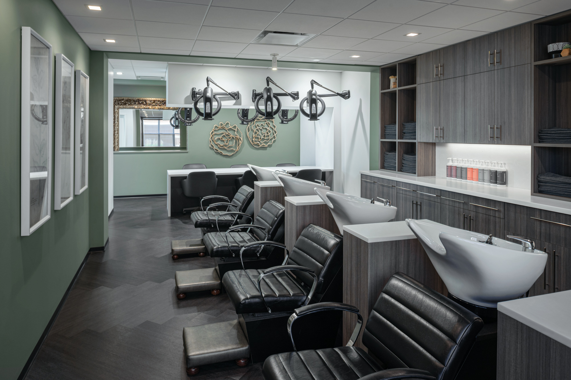 A modern hair salon features sleek black chairs, footrests, and hair-washing stations with white basins. The walls are decorated with abstract art and large mirrors. Dark wood cabinetry houses towels and supplies