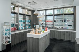 various hair and skincare products neatly arranged on shelves. A central island displays more items, with hanging pendant lights above. The space is bright and well-organized, with large windows letting in natural light.