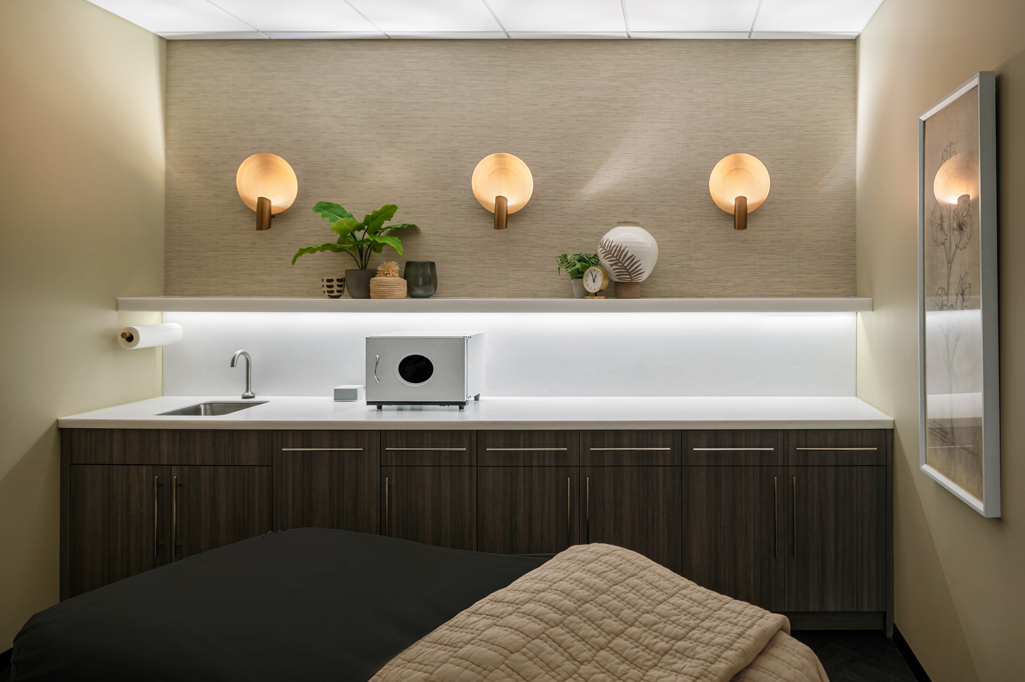 A serene massage therapy room with a massage bed in the foreground covered with a dark blanket and a beige quilt. The background features a white countertop with cabinets, a small sink, plants, and decorative items. Warm lighting fixtures add a cozy ambiance.