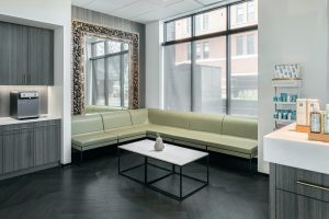 A modern waiting room with large windows, a light green sectional sofa, a white coffee table with a centerpiece, and a framed mirror on the wall.