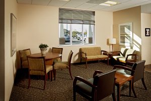 A waiting room with a large window. It has two matching sofas and four armchairs around a wooden coffee table, and a wooden round table with four chairs. A floor lamp stands next to the sofas. The window reveals a parking lot and trees outside.