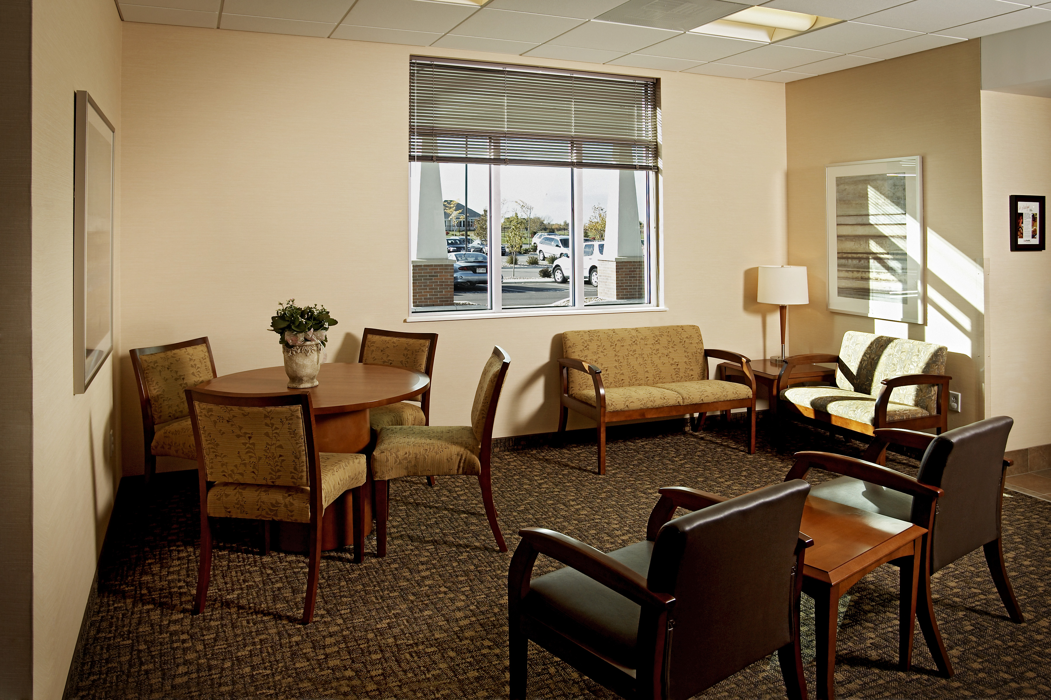 A waiting room with a large window. It has two matching sofas and four armchairs around a wooden coffee table, and a wooden round table with four chairs. A floor lamp stands next to the sofas. The window reveals a parking lot and trees outside.