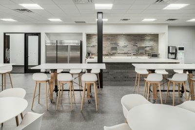 A modern, minimalist kitchen area featuring white tables and stools with light wooden legs. The space has stainless steel refrigerators on the left, a coffee machine and sinks in the back, and a backdrop of grey brick tiles. Walls and ceiling are white.