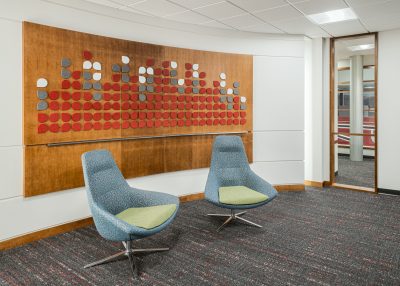 A modern office lobby featuring two blue and green swivel chairs on grey carpet. A decorative wooden panel with red, grey, and white geometric shapes is mounted on the white wall behind the chairs.