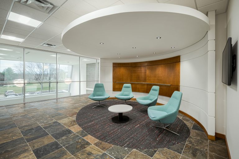 A modern office lounge area with four teal swivel chairs arranged around a small round table on a circular rug. The space has a curved wooden accent wall and large windows offering a view of trees and parked cars. The floor is tiled in a slate pattern.