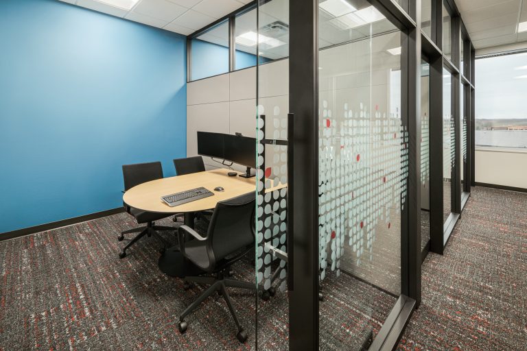 A modern office meeting room with a round wooden table, four black office chairs, and a computer monitor on the table. The room has a blue accent wall, glass partitions with a dotted design, and a carpeted floor in shades of grey and red.