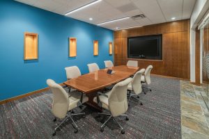 A modern conference room with a long wooden table surrounded by eight beige office chairs. The room has a blue accent wall with three wooden display niches, a large monitor mounted on a wooden panel, and a glass door. The floor has a patterned carpet.